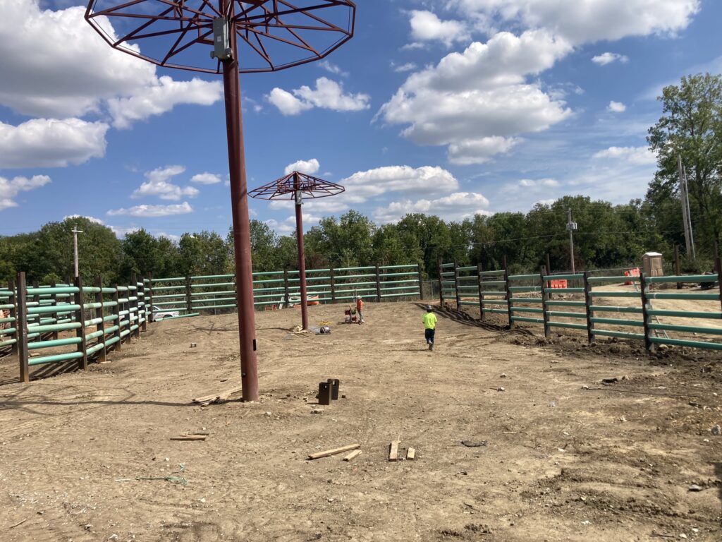 A construction site with two workers and metal structures in a dirt area. Rows of green fencing line the perimeter, with trees and a blue sky with scattered clouds in the background.