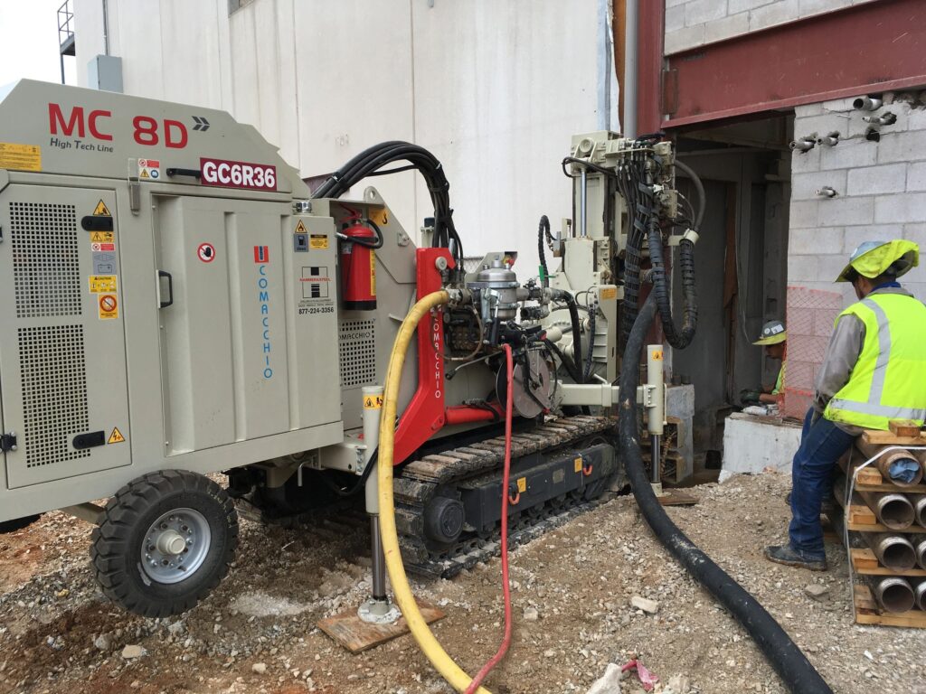 A construction worker wearing a neon vest and helmet operates a large drilling machine near a building wall. The machine has various hoses and cables attached, with a stack of pipes nearby on the ground.