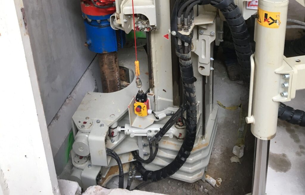 A close-up of industrial machinery, featuring a drill and various components such as cables, rods, and gears. The equipment appears to be set up against a wall in a construction or manufacturing environment.