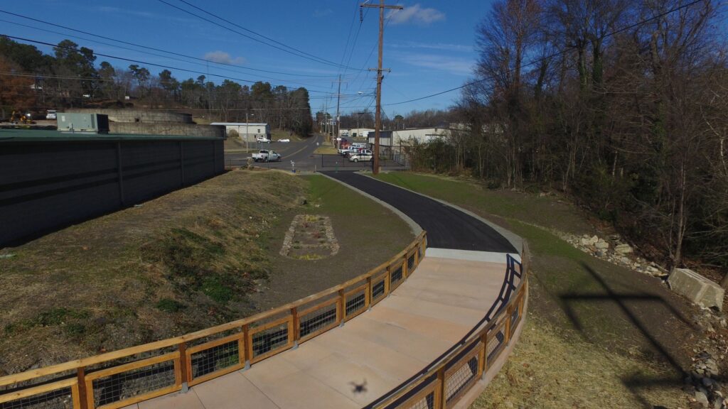 A paved path curves alongside a street, bordered by a wooden railing. The area has sparse grass and trees, utility poles, and buildings in the distance. The sky is clear and blue.