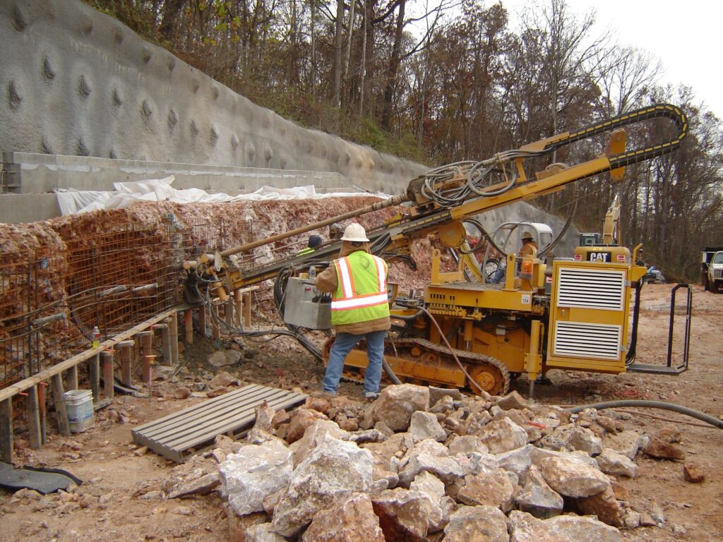 A worker in a safety vest and helmet operates heavy machinery at a construction site, drilling into a rocky surface. The area is surrounded by loose stones, dirt, and a concrete wall. Trees and equipment are visible in the background.