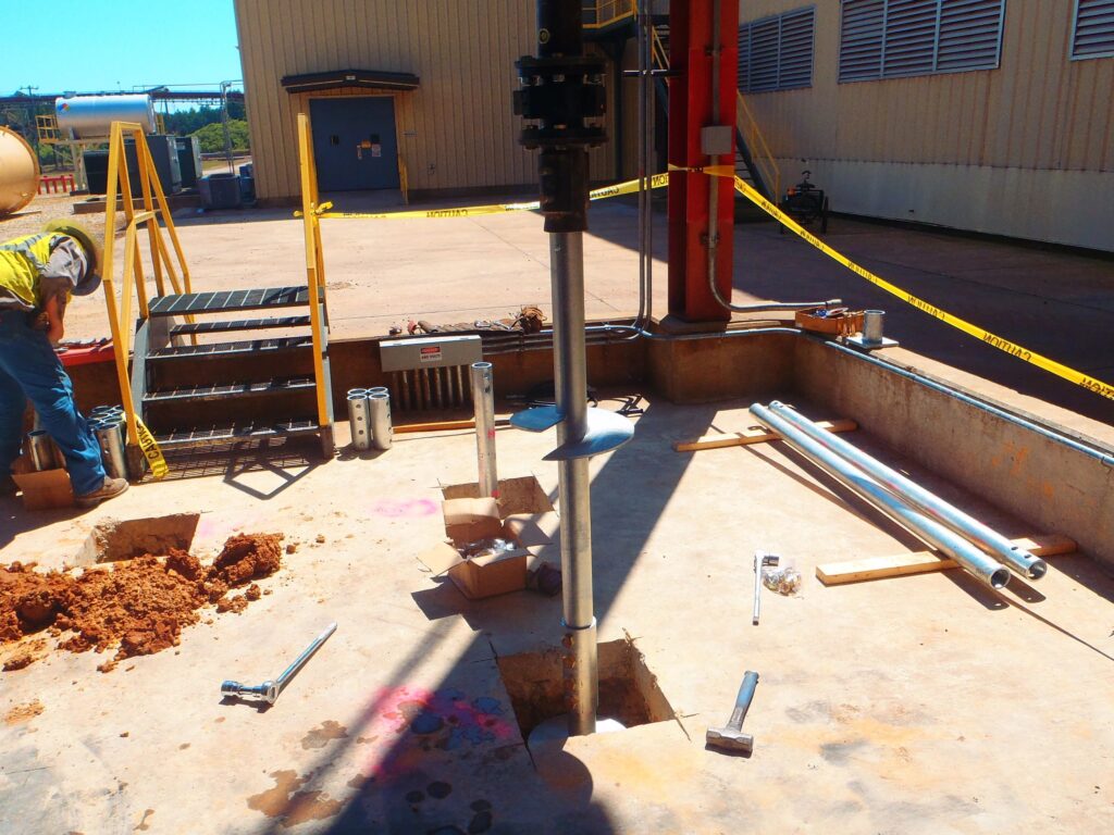 A construction site with a worker installing a metal post into a hole. Surrounding tools include wrenches and pipes. Yellow caution tape is in the background, along with a building and other industrial equipment.