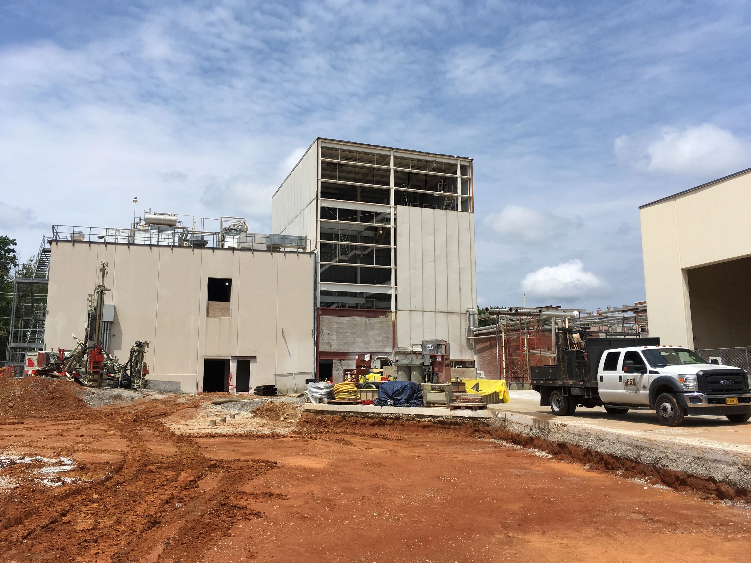 Construction site with a partially constructed industrial building framed by blue sky. Construction materials and vehicles, including a white truck and drilling machinery, are present around the site. Earth is excavated in the foreground.