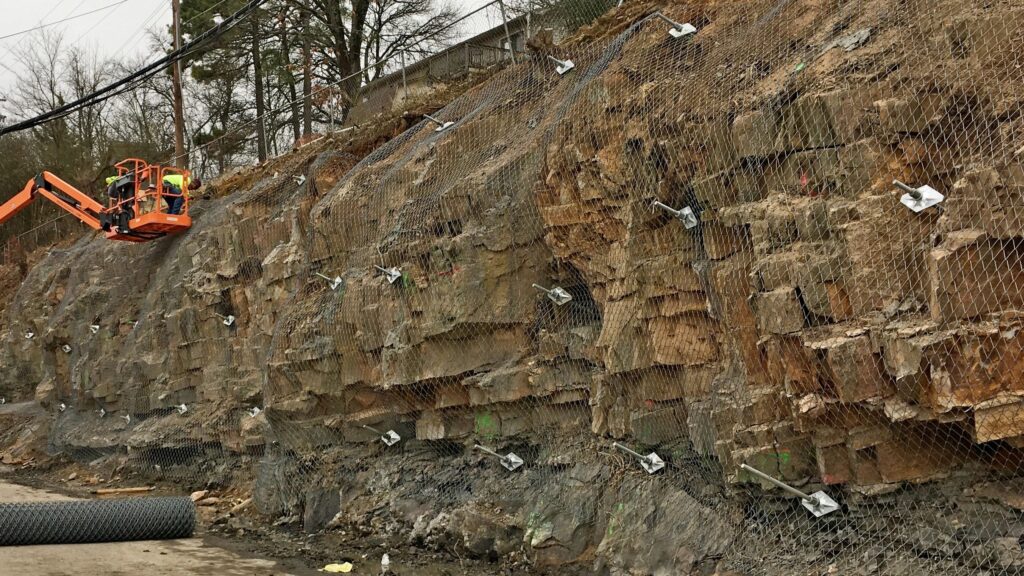 Construction workers in an orange lift secure wire netting to a rocky cliff face. The netting is fastened with metal anchors. Nearby, a roll of material is seen on the ground, with trees and utility lines in the background.