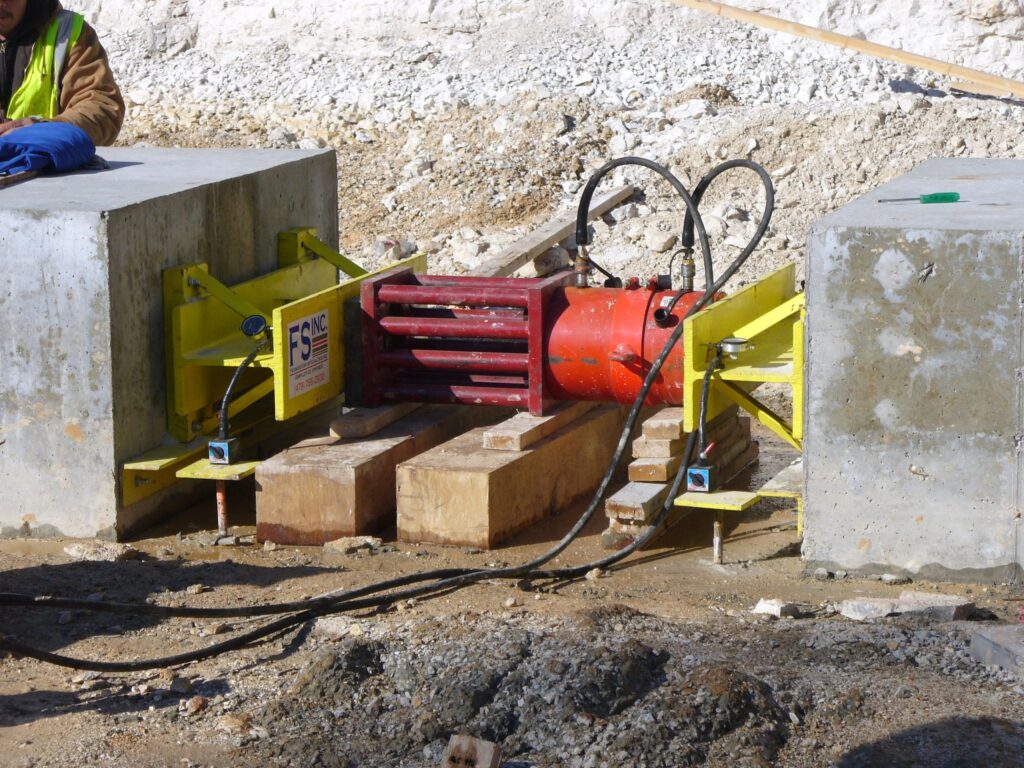 A construction site featuring a hydraulic jacking system positioned between two concrete blocks. The system includes a red hydraulic cylinder and yellow framework. The ground is bare and surrounded by gravel and sand. A worker is partially visible on the left.