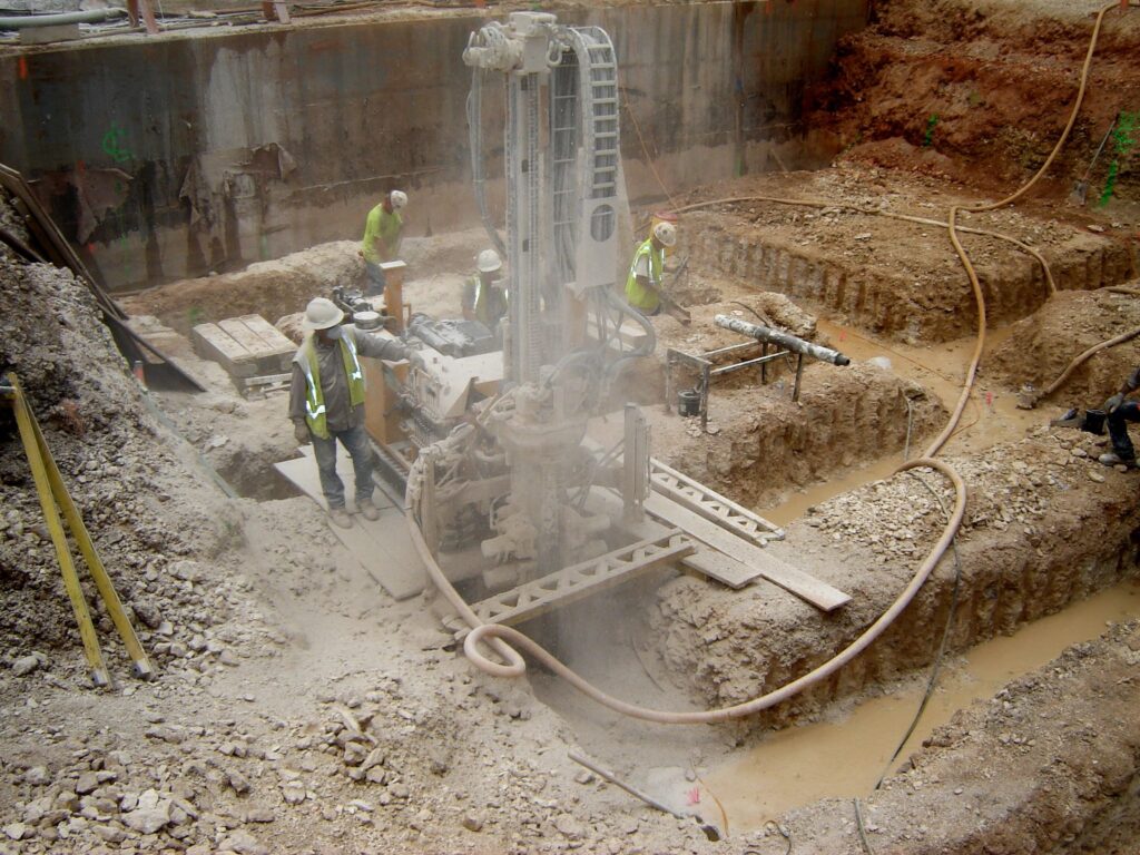 Construction workers operate a drilling machine in a muddy excavation site. They are wearing safety gear, including helmets and vests. The site is surrounded by dirt and construction materials.