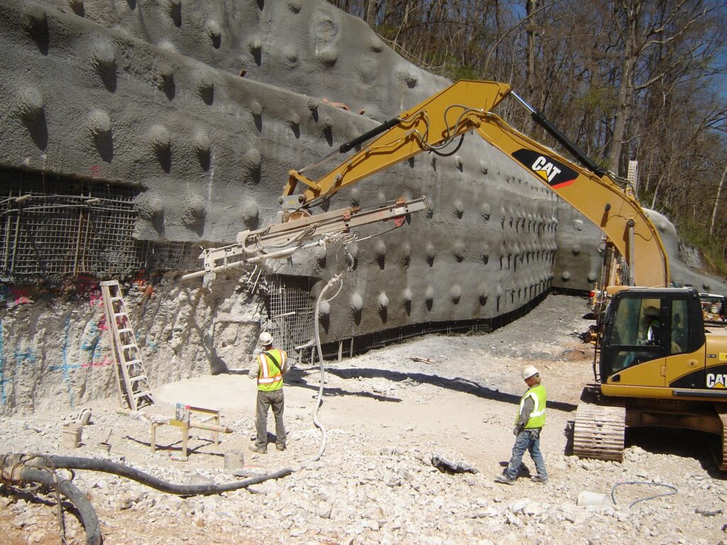 Construction workers operate heavy machinery, including an excavator and a drilling rig, against a textured concrete wall. The ground is covered with gravel and equipment. Workers wear safety gear, including hard hats and vests. Trees are visible behind.