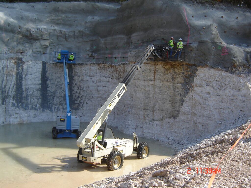 A construction site with two elevated work platforms. Workers in safety gear are operating equipment along a rocky wall. The ground is partially covered with water, and there are markings on the rock face.