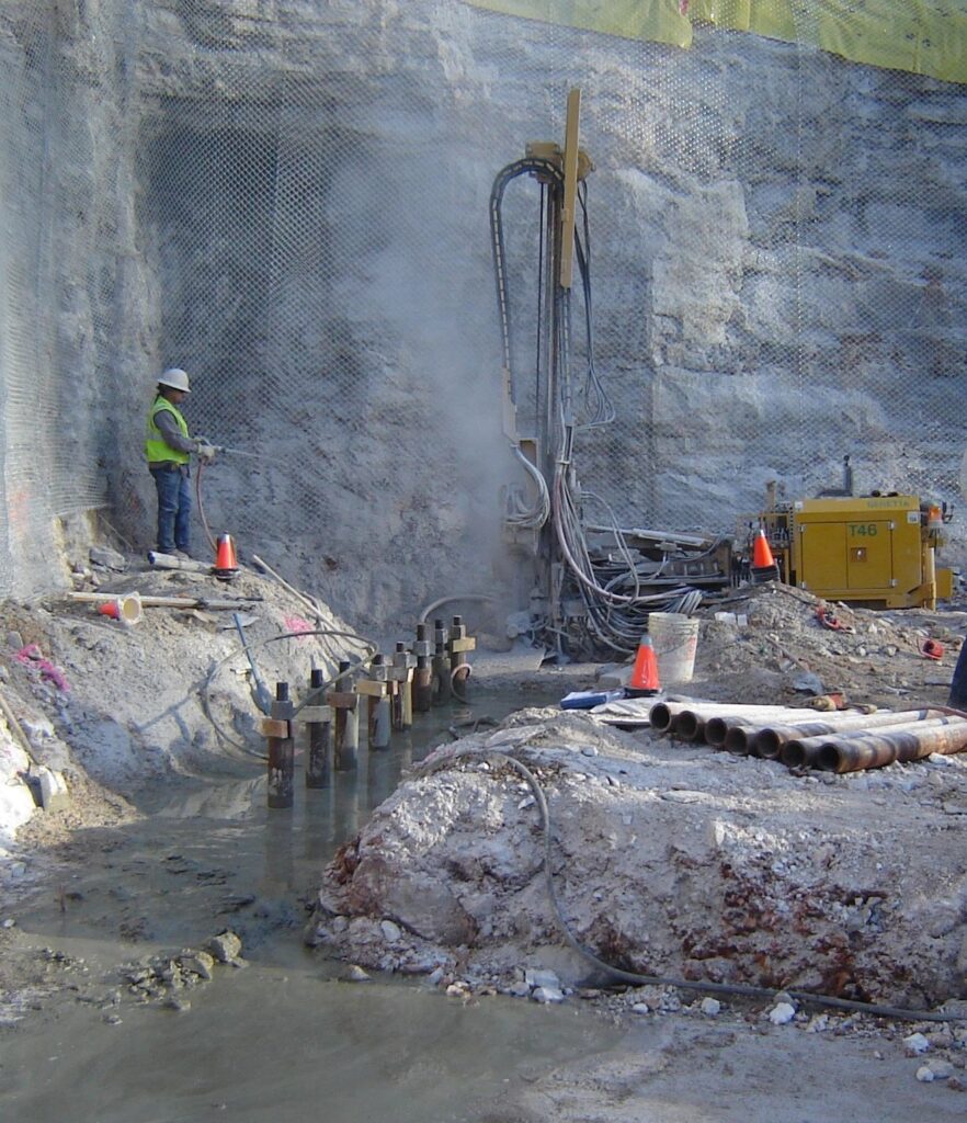 Construction site with a worker in a safety vest and helmet operating machinery near a rocky wall. Visible pipes and wet ground surround the area, with orange cones marking the site. A yellow machine is situated in the background.