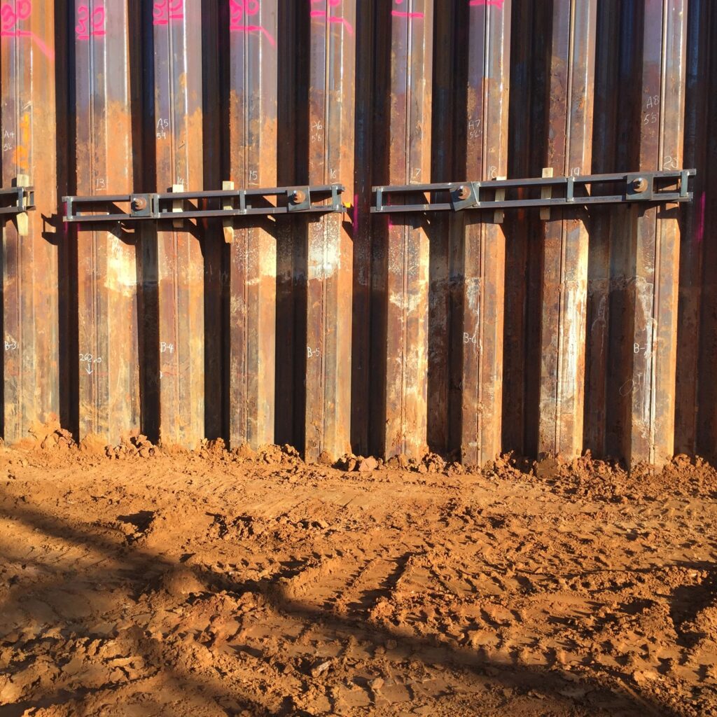 Wall of rusted metal beams with horizontal metal brackets near the top, set against a backdrop of sunlit, loose red soil in a construction area. Pink markings are visible at the top of the beams. Shadows stretch across the ground.