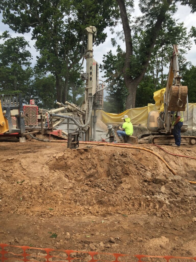 Two construction workers operate heavy machinery at a construction site surrounded by trees. One worker sits next to a drill, while another stands nearby. The area is marked off with an orange safety fence.