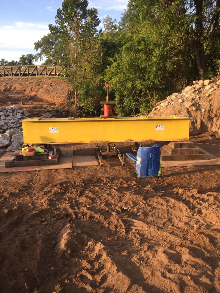 A construction site with a yellow industrial machine on a sandy ground. There are trees and a bridge in the background. Some tools and equipment are placed nearby.