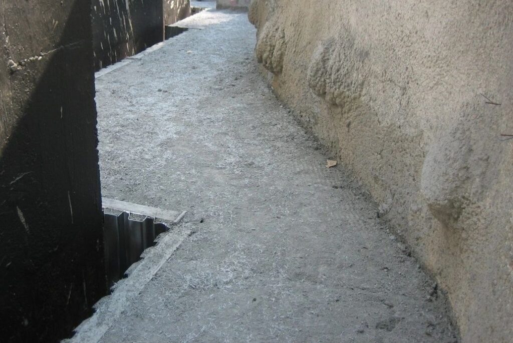 A narrow, concrete alleyway with uneven surfaces is bordered by high walls. The path curves slightly, disappearing into the background and showing a metal grate on the left side. Shadows from the walls create a contrast with the light gray walkway.