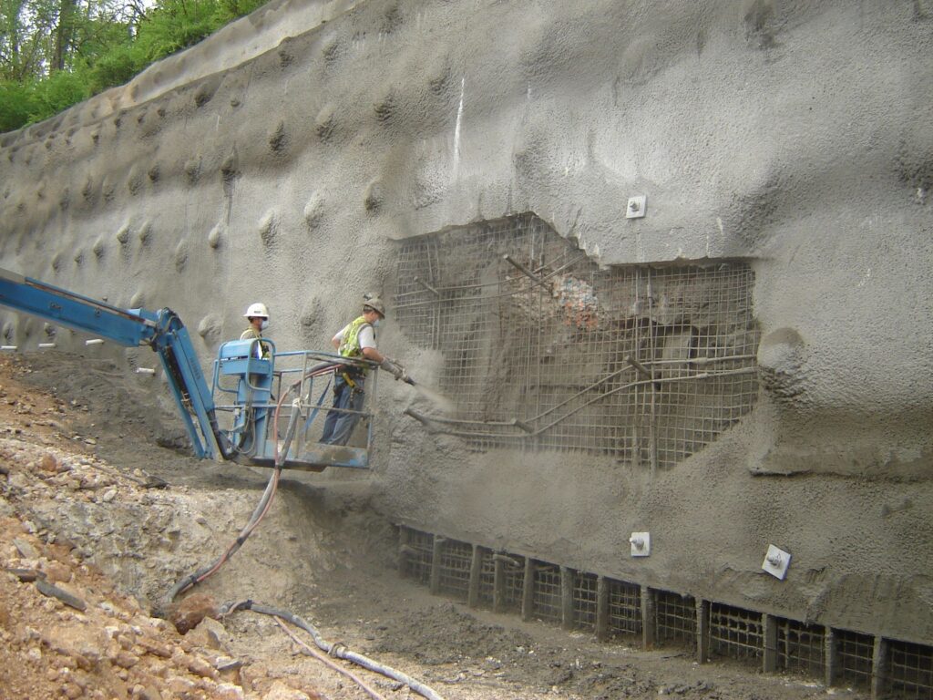 Two construction workers in safety gear operate a hose on a blue lift, spraying concrete onto a reinforced wall. The wall has exposed rebar and a rough surface, with a partially filled section. Dirt and construction debris are visible on the ground.