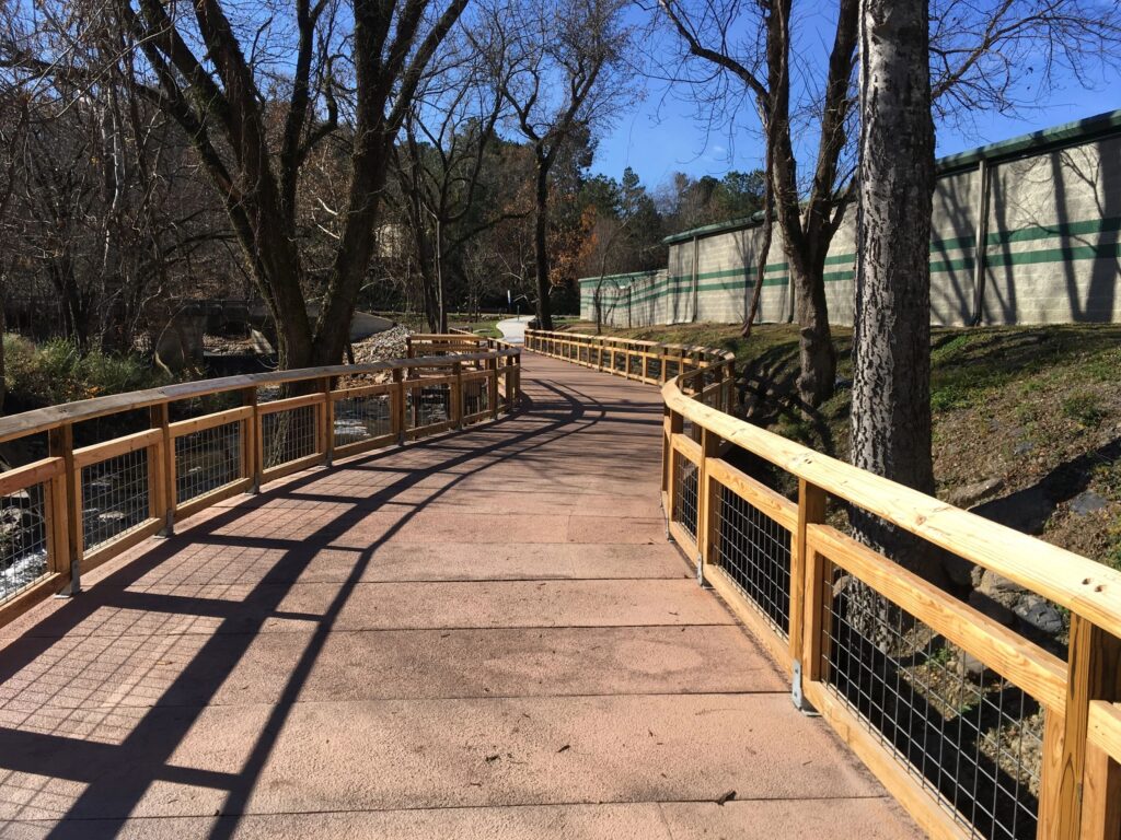 A paved pathway winds through a park, bordered by wooden railings and trees. The blue sky above indicates a clear, sunny day. The path curves gently to the left, leading into a wooded area.