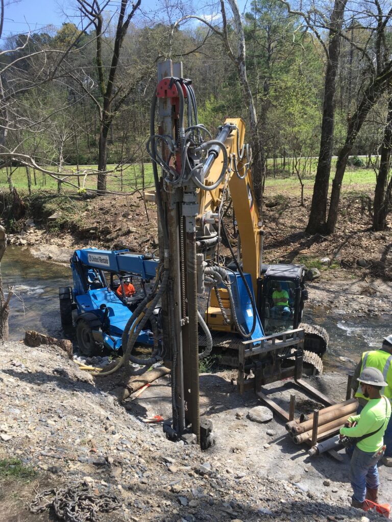 A construction site near a small creek with workers operating heavy machinery, including a drilling rig and a blue utility vehicle. Two workers in helmets and vests are working with large wooden beams. Trees and a grassy area are visible in the background.