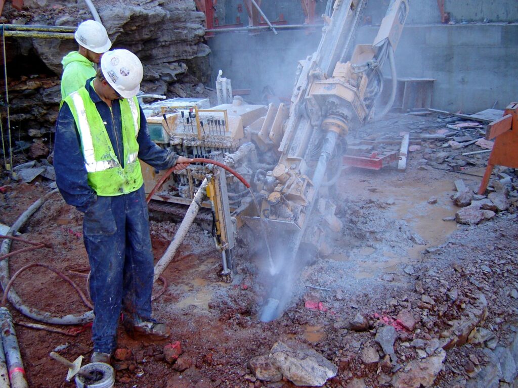 Two construction workers in hard hats and safety vests operate a large, drilling machine at a rocky worksite. One worker uses a hose to manage dust or debris, while the other oversees the equipment. Dust and water create a visible mist around them.