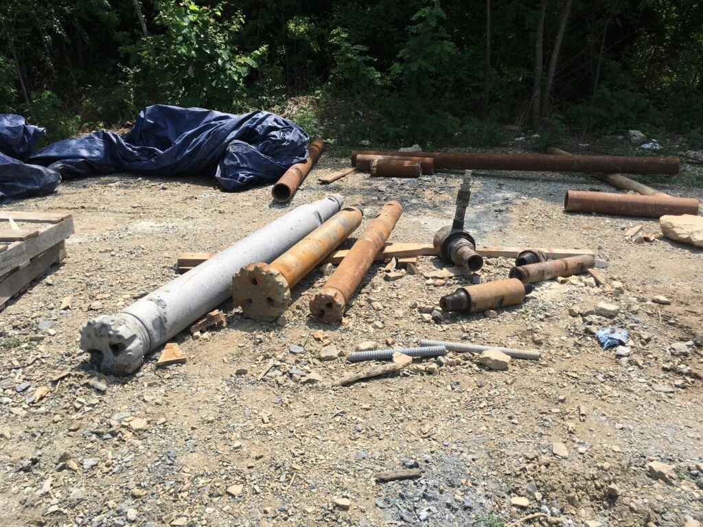 Several rusted metal pipes and cylindrical objects are scattered on a gravel surface, surrounded by some trees and a tarp on the left. The area appears to be an outdoor construction or storage site.