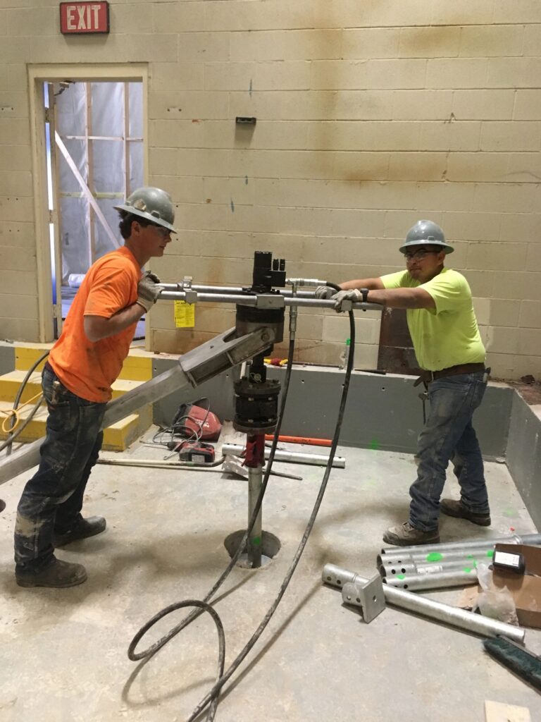 Two construction workers in hard hats and safety gear operate a metal pipe cutter inside a building. One man wears an orange shirt, and the other wears a yellow shirt. They are standing on a concrete floor with tools and materials around. A door marked "EXIT" is in the background.