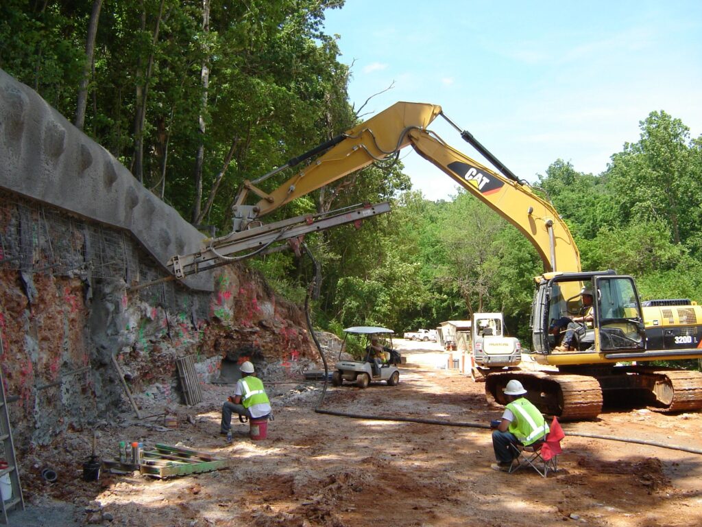 Workers in safety gear operate machinery on a construction site with a large excavator digging into a rocky hillside. One worker uses a tool on the ground, while another observes. The site is surrounded by dense greenery under a clear sky.