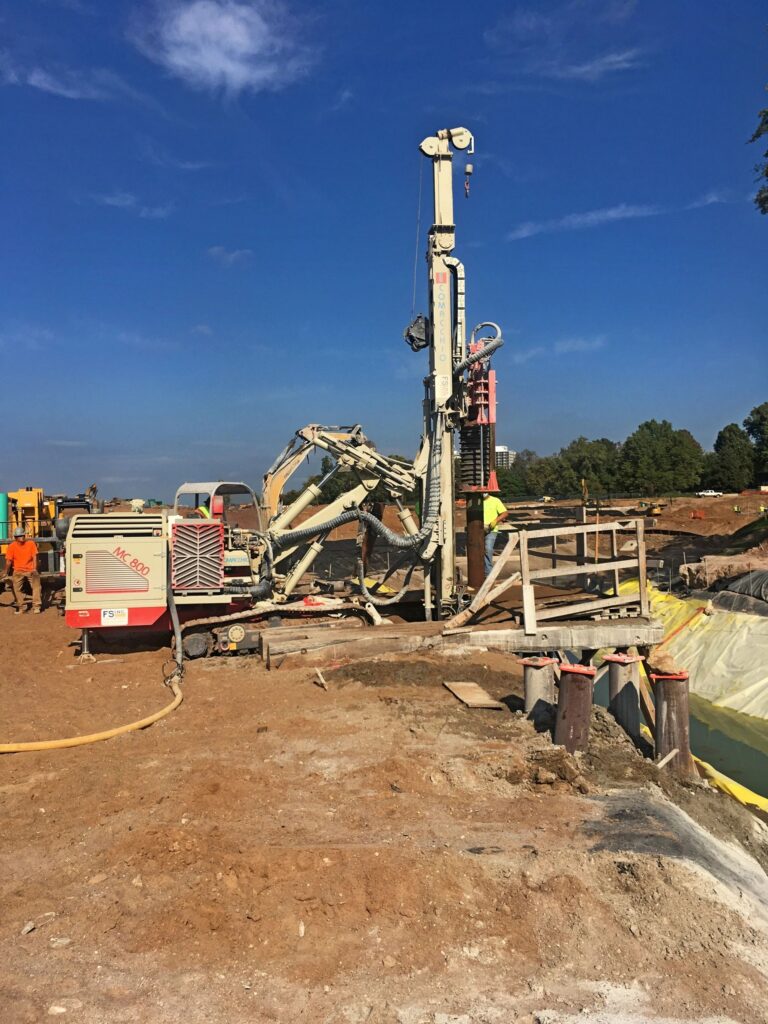 A construction site with a drilling machine in operation on a clear day. Workers in safety gear are managing the equipment, surrounded by dirt and machinery. In the background, there are trees and open land.