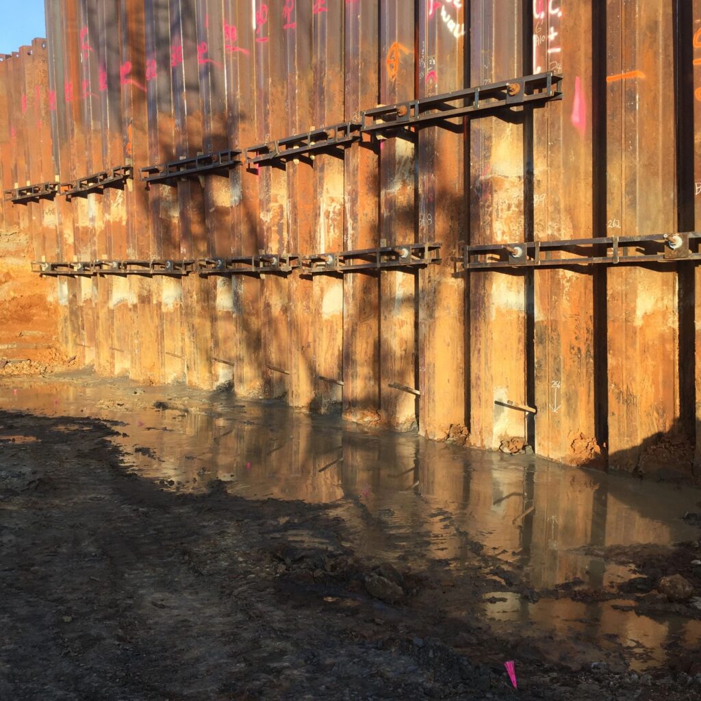 A metal retaining wall at a construction site, reinforced with beams and surrounded by muddy ground. Sunlight casts shadows on the wall, highlighting its structure.