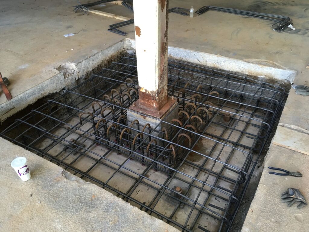 A construction site showing a concrete foundation with exposed steel reinforcement bars surrounding a vertical metal column. Tools and a small container are nearby, with the area partially excavated.