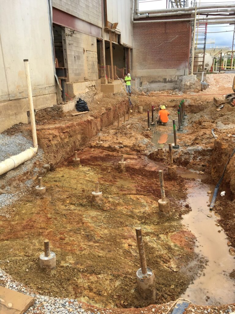 Construction site with exposed earth, several metal rods protruding from the ground, and a partially demolished building wall. Two workers in high-visibility vests and helmets are on site. Puddles of water are visible around the area.