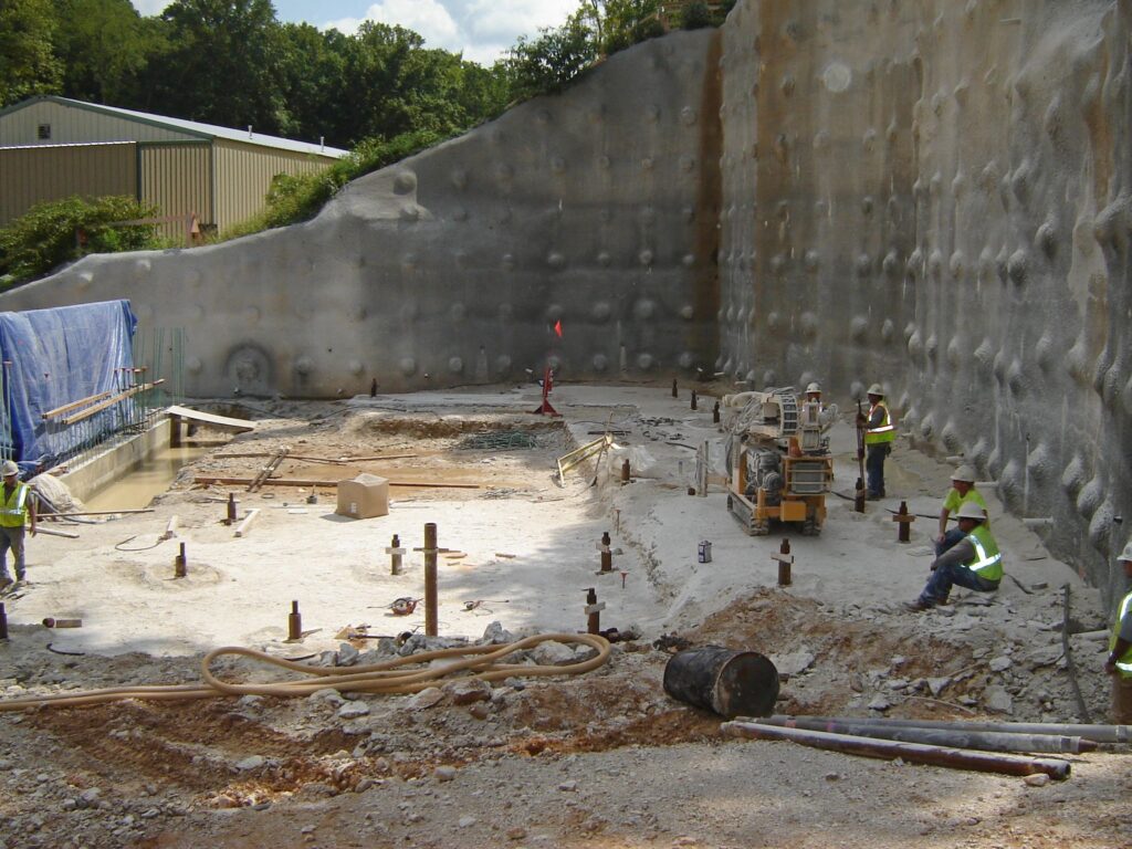 Construction site with workers wearing high-visibility clothing and helmets. They are preparing ground for a building foundation, surrounded by a tall, reinforced wall. Tools, machinery, and materials are scattered around the area.