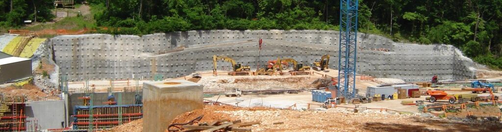 A construction site with several excavators working near a large concrete retaining wall. There is a blue crane tower, various construction materials, and forested areas in the background.