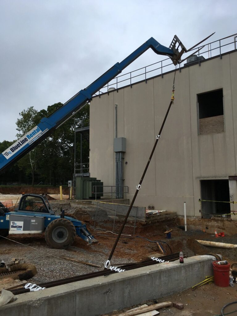 A construction site with a blue crane lifting a long metal beam into place on a building structure. The crane is positioned on a rocky, dirt-covered surface next to a concrete wall. Safety barriers and equipment are visible on the ground.