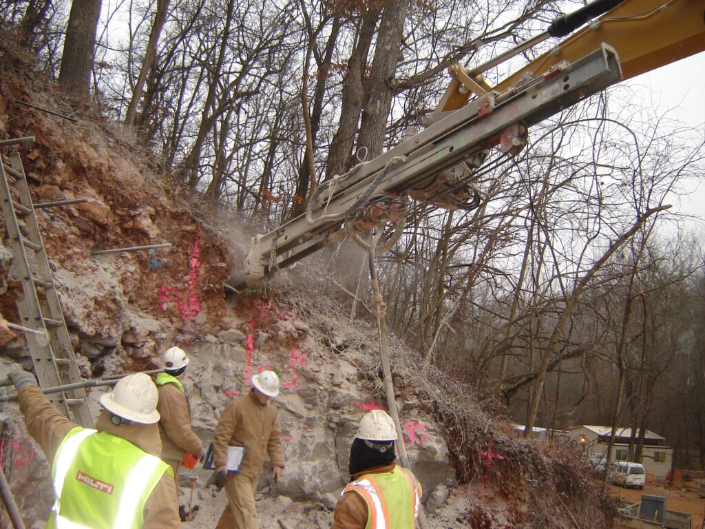 Construction workers operating heavy machinery to drill into a rocky hillside. They are wearing safety vests and helmets. The area is surrounded by trees, and there are visible pink markings on the rocks.