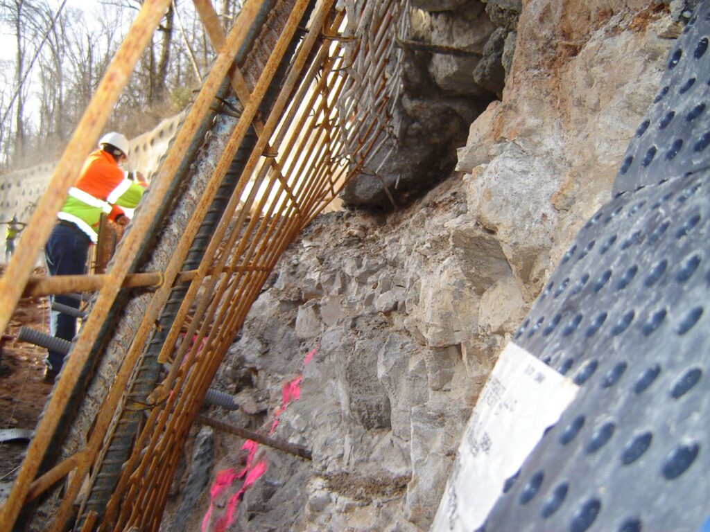 A construction worker in safety gear stands by a rugged rock slope reinforced with metal rods and grating. Bright pink markings are visible on the rocks, and a black tarp covers part of the slope. Trees can be seen in the background.