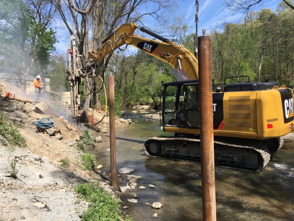 A construction site by a stream, featuring a yellow excavator and workers in safety gear. Two tall metal poles are being installed on the rocky bank. Lush trees surround the area, and sunlight filters through the branches.