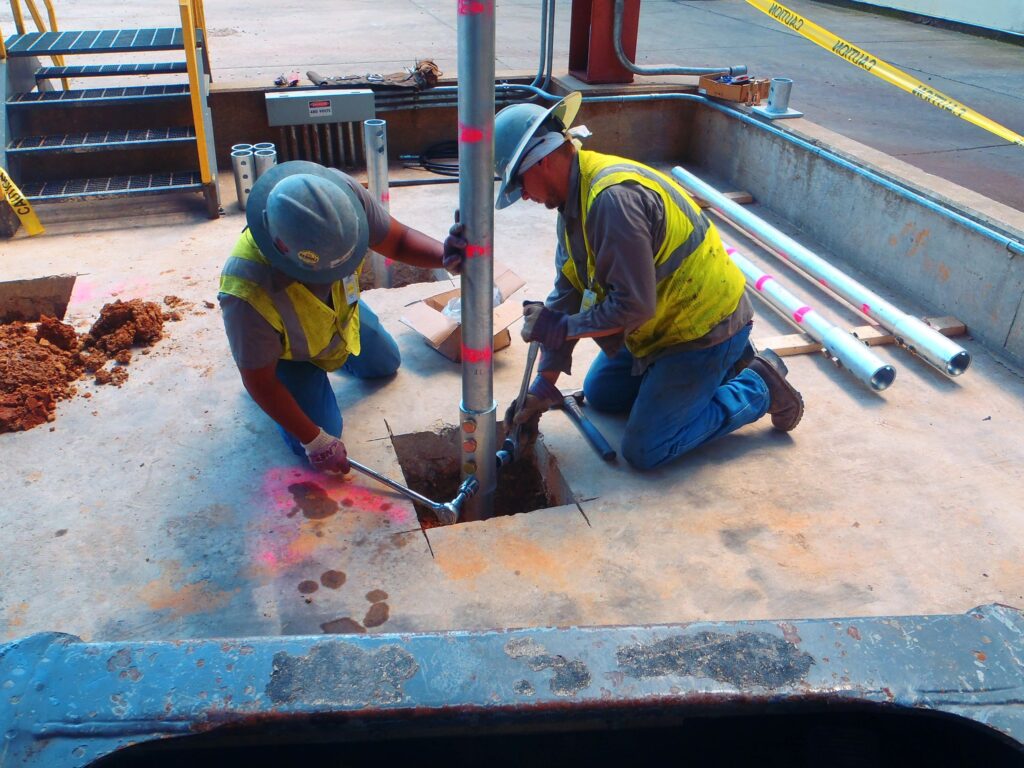 Two construction workers in safety gear are installing a metal pole in a concrete floor. They are using tools to secure the pole in place. The area is marked with caution tape and construction equipment is visible around them.