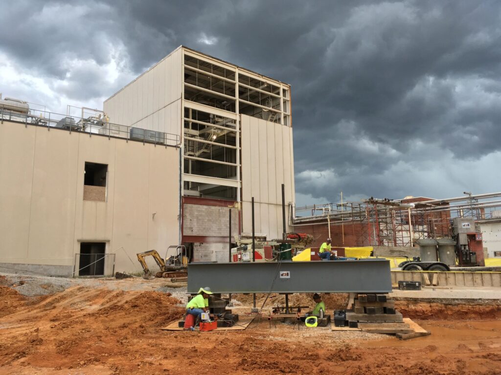 Construction site with workers preparing infrastructure near a large industrial building. The sky is overcast with dark clouds, indicating an approaching storm. Heavy machinery and building materials are visible around the area.