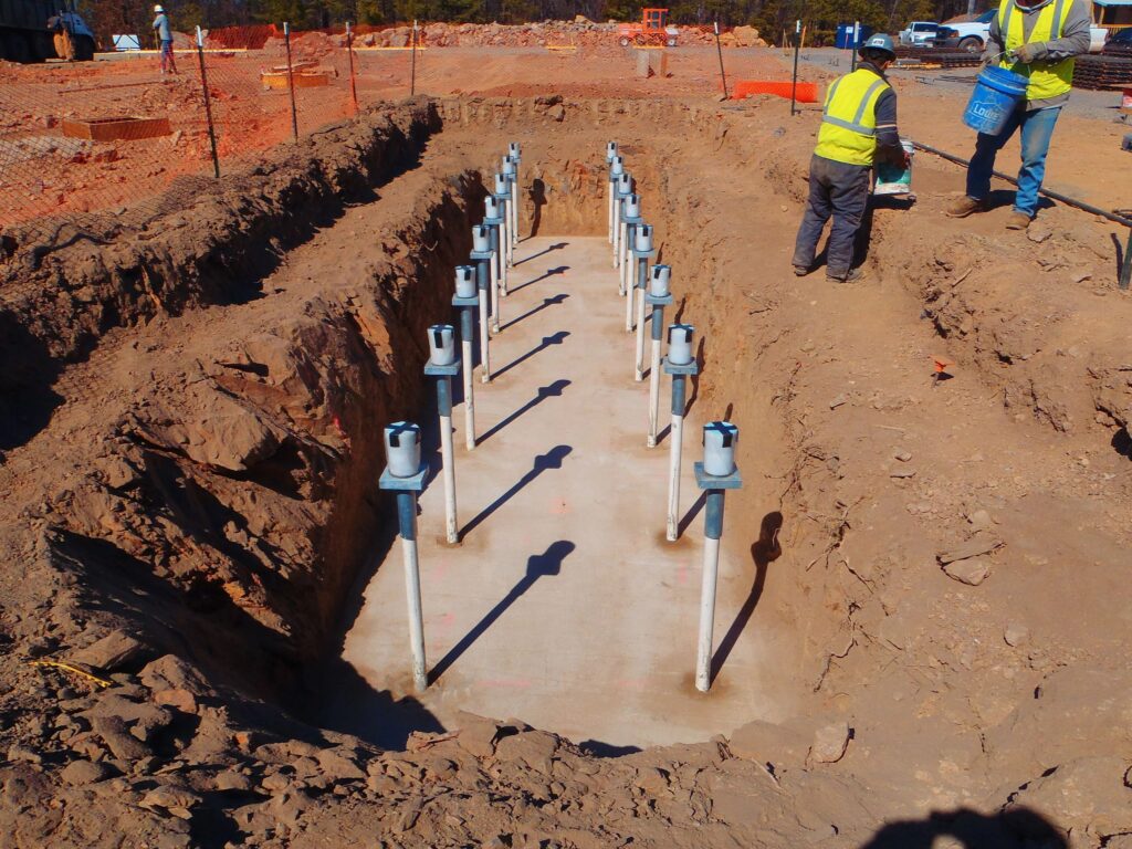 A construction site showing a rectangular trench with multiple vertical pipes protruding from the concrete base. Two workers in safety gear stand near the edge of the trench, surrounded by soil and orange safety netting.