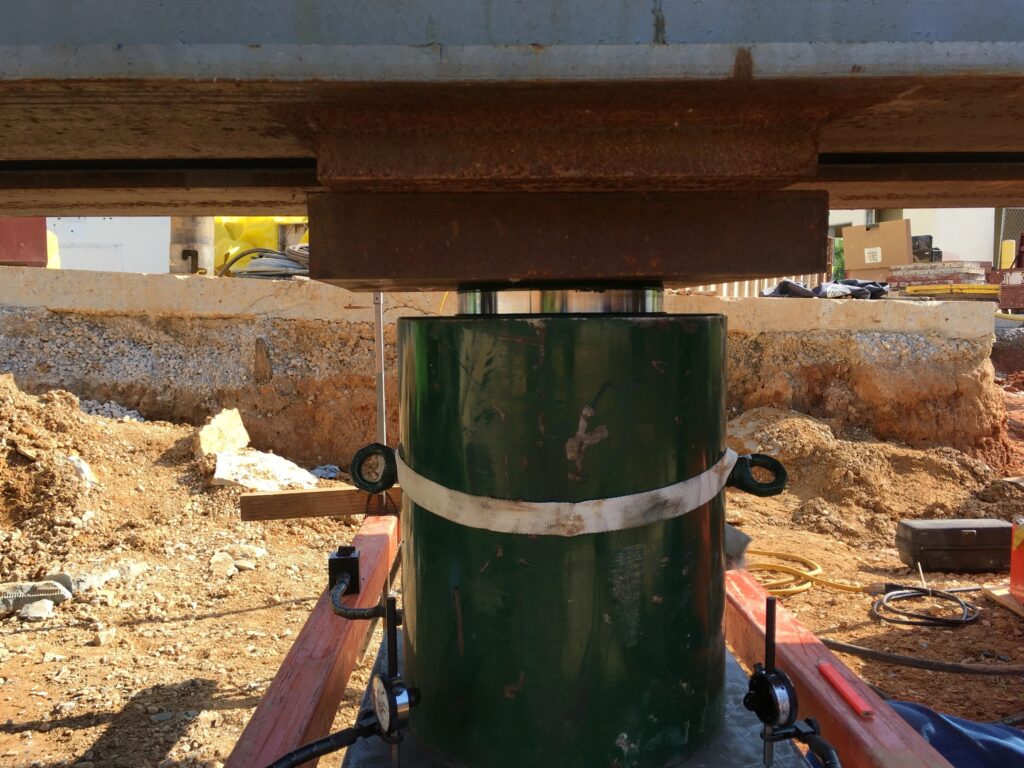 A hydraulic jack supports a large metal beam at a construction site. The jack is green and surrounded by a metal frame. The background shows soil, construction materials, and tools scattered around.