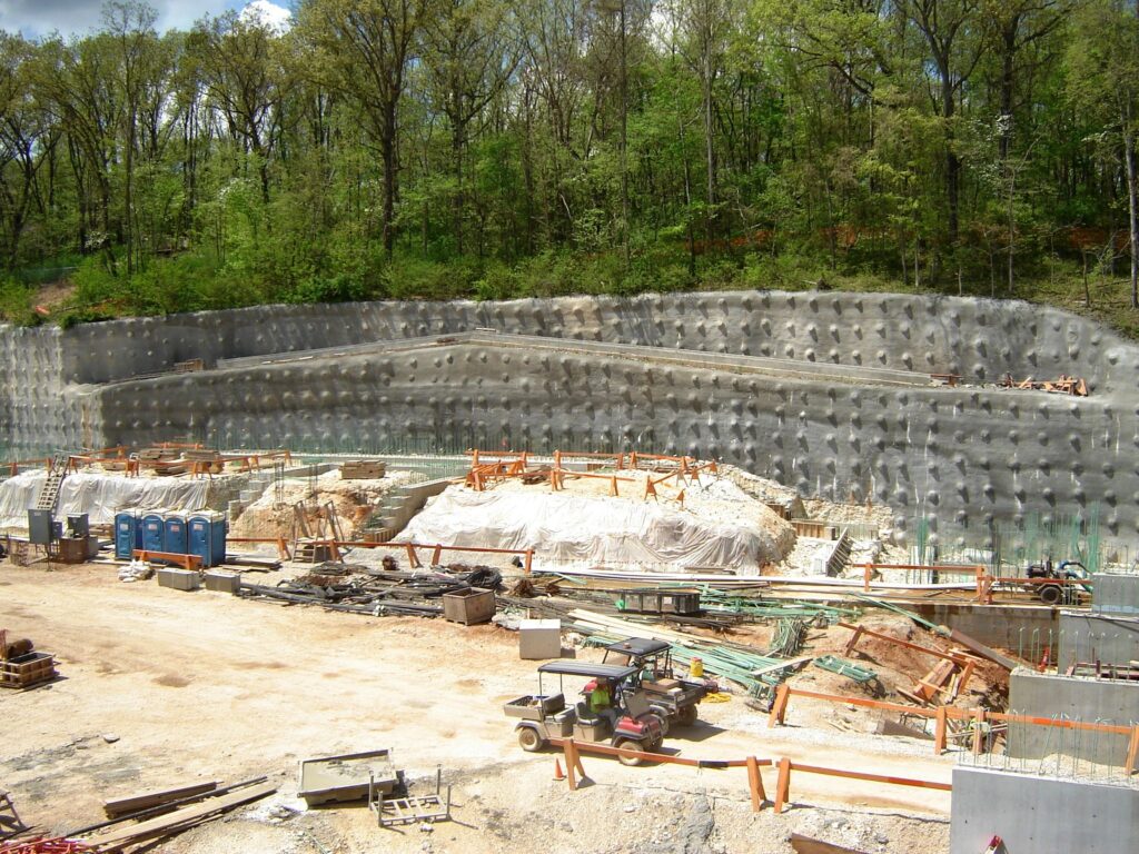 A construction site surrounded by trees features a partially completed retaining wall with a stepped design. Equipment and materials are scattered around the area, and a row of portable toilets is visible on the left.