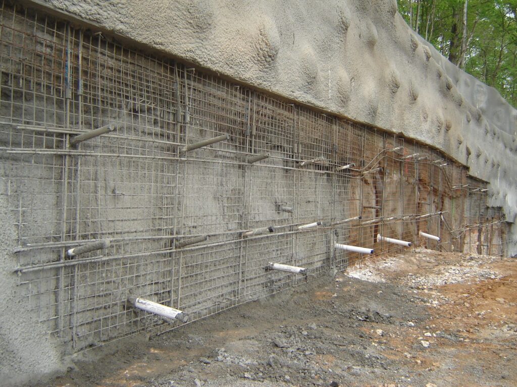 A reinforced concrete wall under construction, with exposed steel rebar and wooden beams protruding from the surface. The surrounding ground is bare soil, and there are trees in the background.