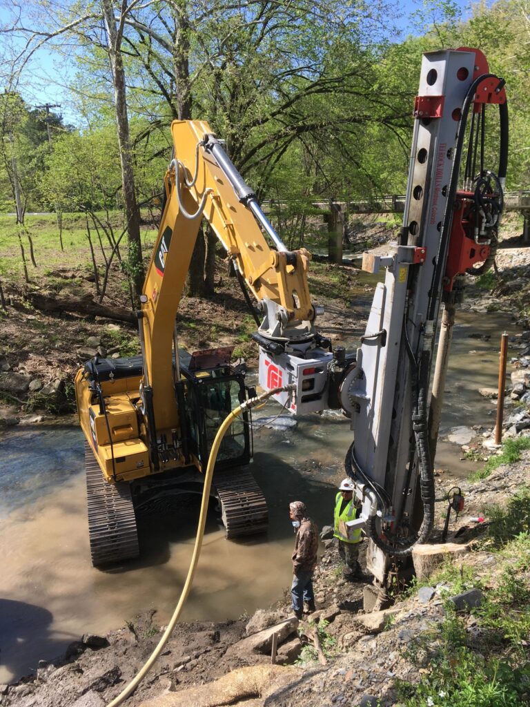 A yellow excavator operates near a small river, positioned next to a vertical drilling rig. Two workers stand nearby, overseeing the equipment. Surrounding the scene are trees and natural vegetation under a clear blue sky.