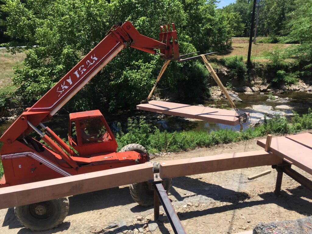 A red telescopic handler is lifting a wooden plank over a small stream, surrounded by green trees and grass. The machine, labeled "SkyTrak," is part of a construction project near the water's edge.