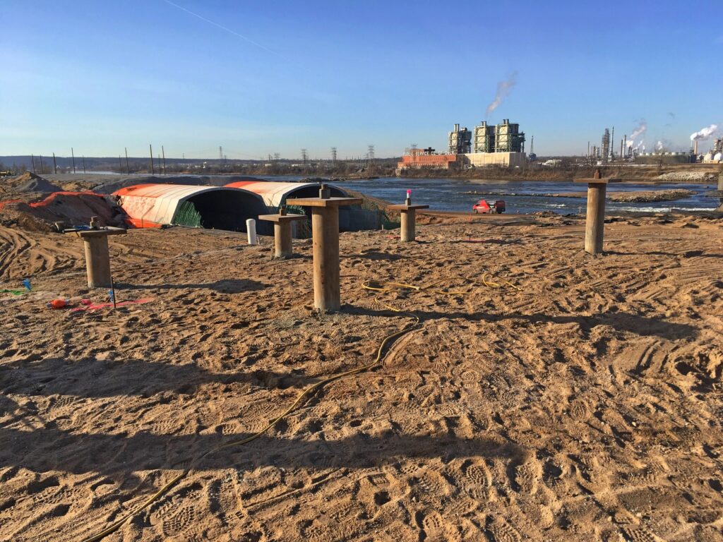 A sandy construction site with wooden posts, cables, and covered structures in the foreground. An industrial facility with smokestacks is visible in the background, next to a body of water under a clear blue sky.