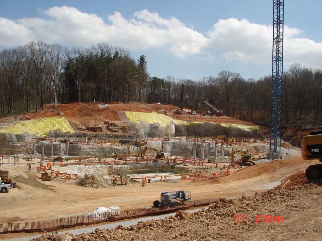 Construction site in a wooded area with exposed rock walls, heavy machinery, and cranes. Earth movers and vehicles are seen within a large excavation area. Yellow tarps cover parts of the hillside, and the sky is partly cloudy.