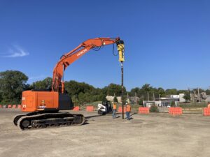 An orange excavator with an attached drill stands on a construction site. Two workers in helmets observe nearby. Orange barriers line the area, and trees are visible in the background under a clear blue sky.