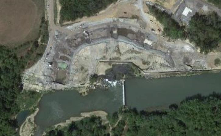 Aerial view of a construction site near a river, featuring a bridge or dam structure. The area is surrounded by dense green trees, and there are pathways and cleared patches of land visible around the site.