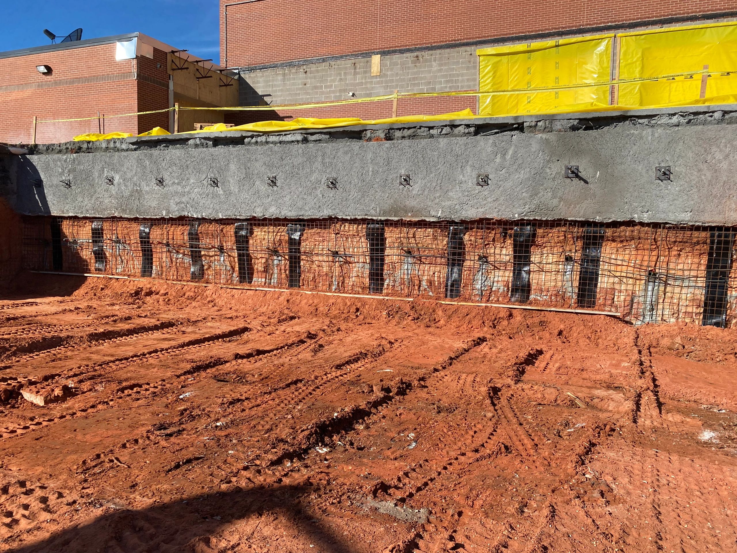 Construction site with exposed foundation wall partially covered in concrete. Yellow tarps hang above, supported by a brick building. The ground is reddish-brown soil with visible tire tracks from construction vehicles. Bright, clear day.