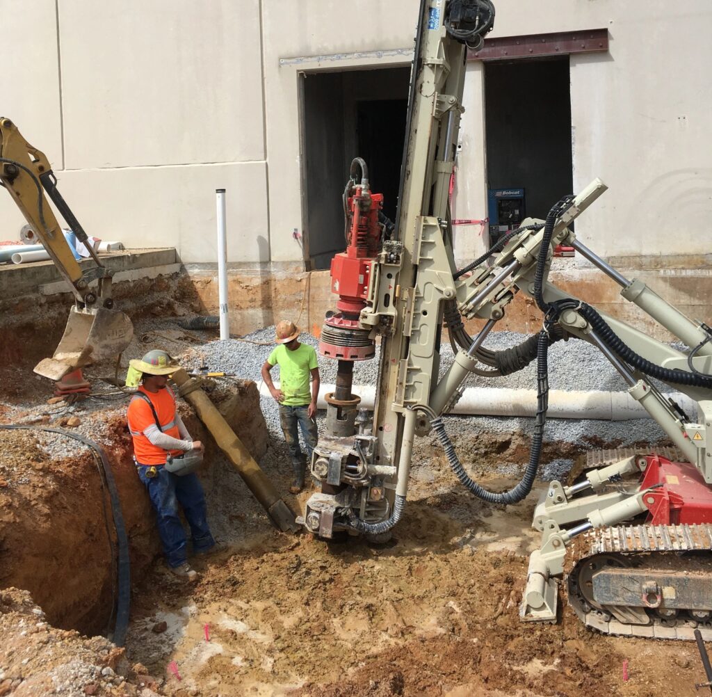 Construction workers operating heavy machinery at a building site. They are drilling into the ground. One worker wears an orange vest and straw hat, another in a yellow vest. There's a partially dug hole and a large building wall in the background.