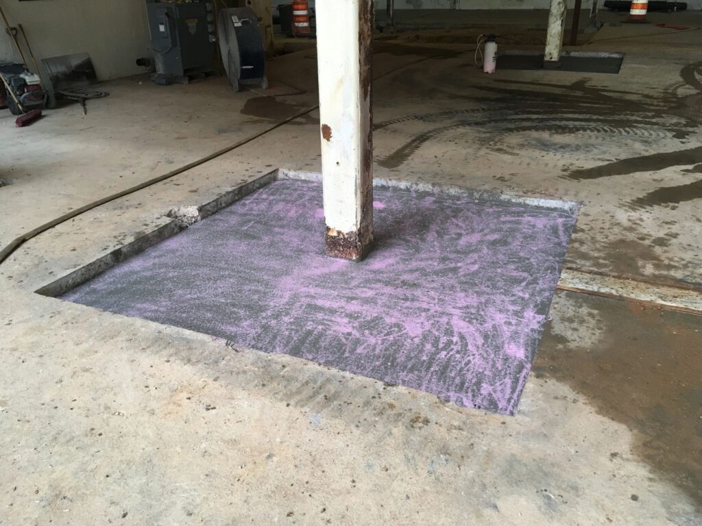 A construction site with a square section of the floor removed, revealing a wooden post embedded in a pink and gray concrete mixture. The site appears to be in a large indoor space with various equipment and construction materials around.