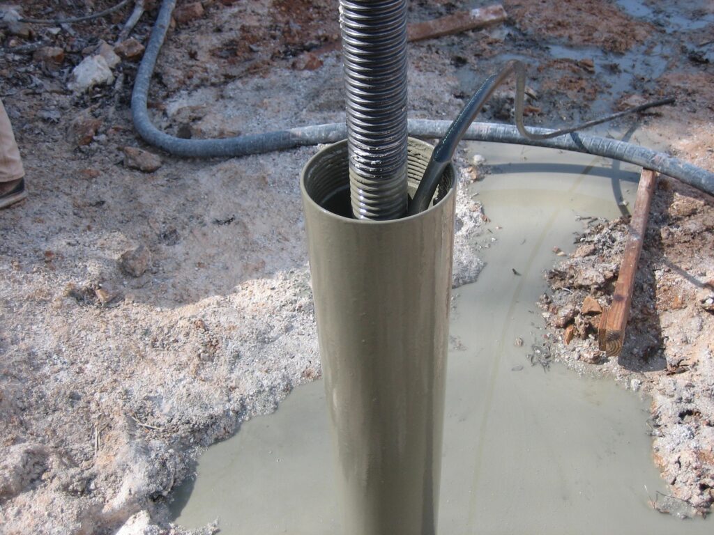 A large pipe is partially embedded in the ground with a flexible hose inside. The surrounding area is muddy and wet, with other hoses and debris visible on the ground.